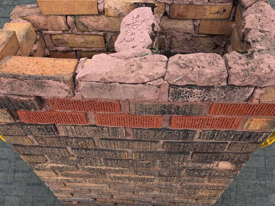The top of a brick chimney with crumbling, washed-out mortar and eroded brick where the crown has failed.