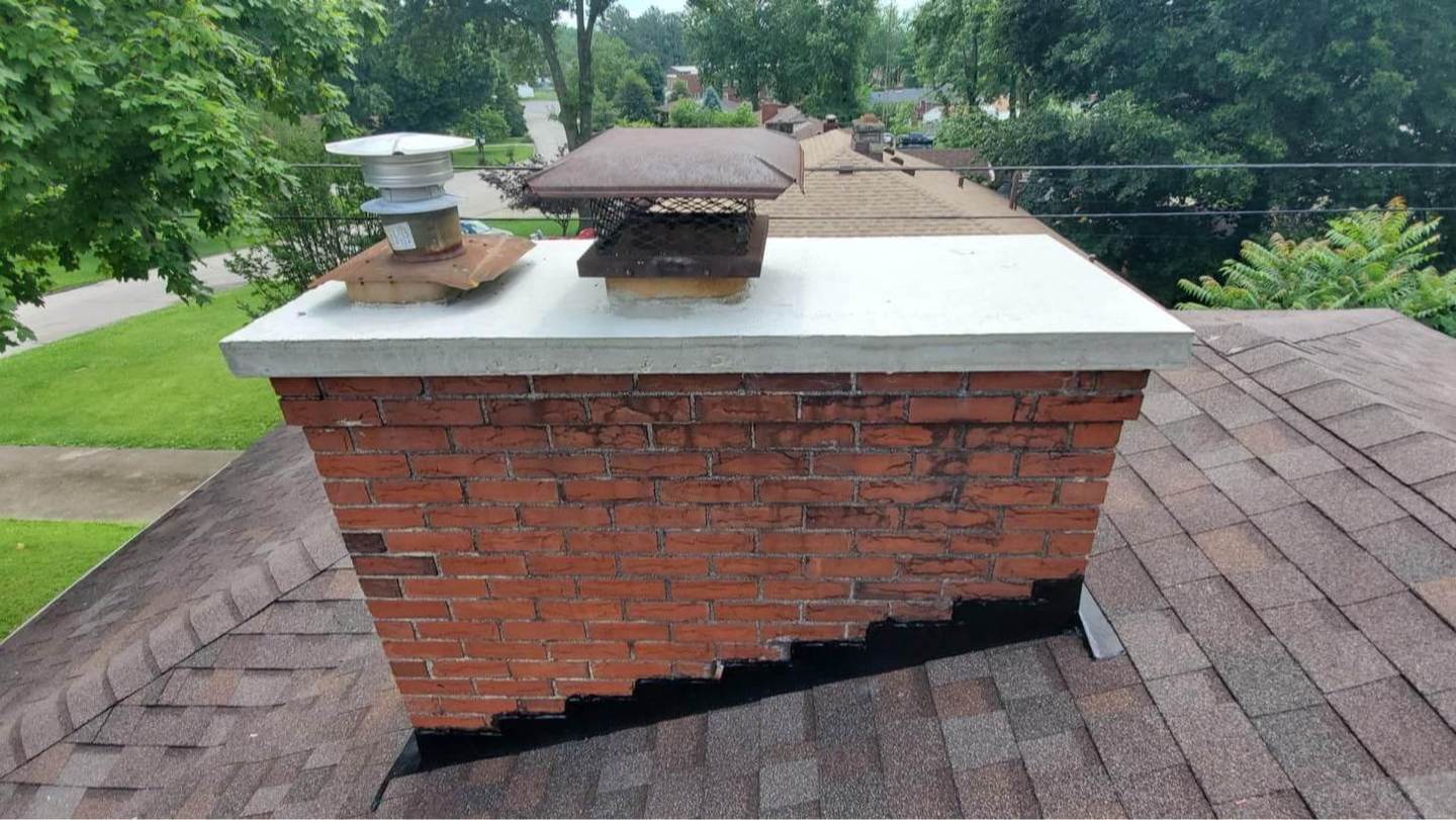 A wide brick chimney topped with a freshly finished concrete crown and two dark metal caps, seen from the shingle roof with a tree-lined street behind it.