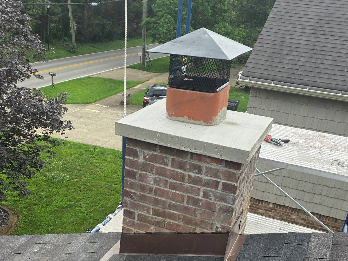 A brick chimney chase with a fresh concrete crown and a black spark-arrestor cap, photographed from the roof with the front lawn and street below.