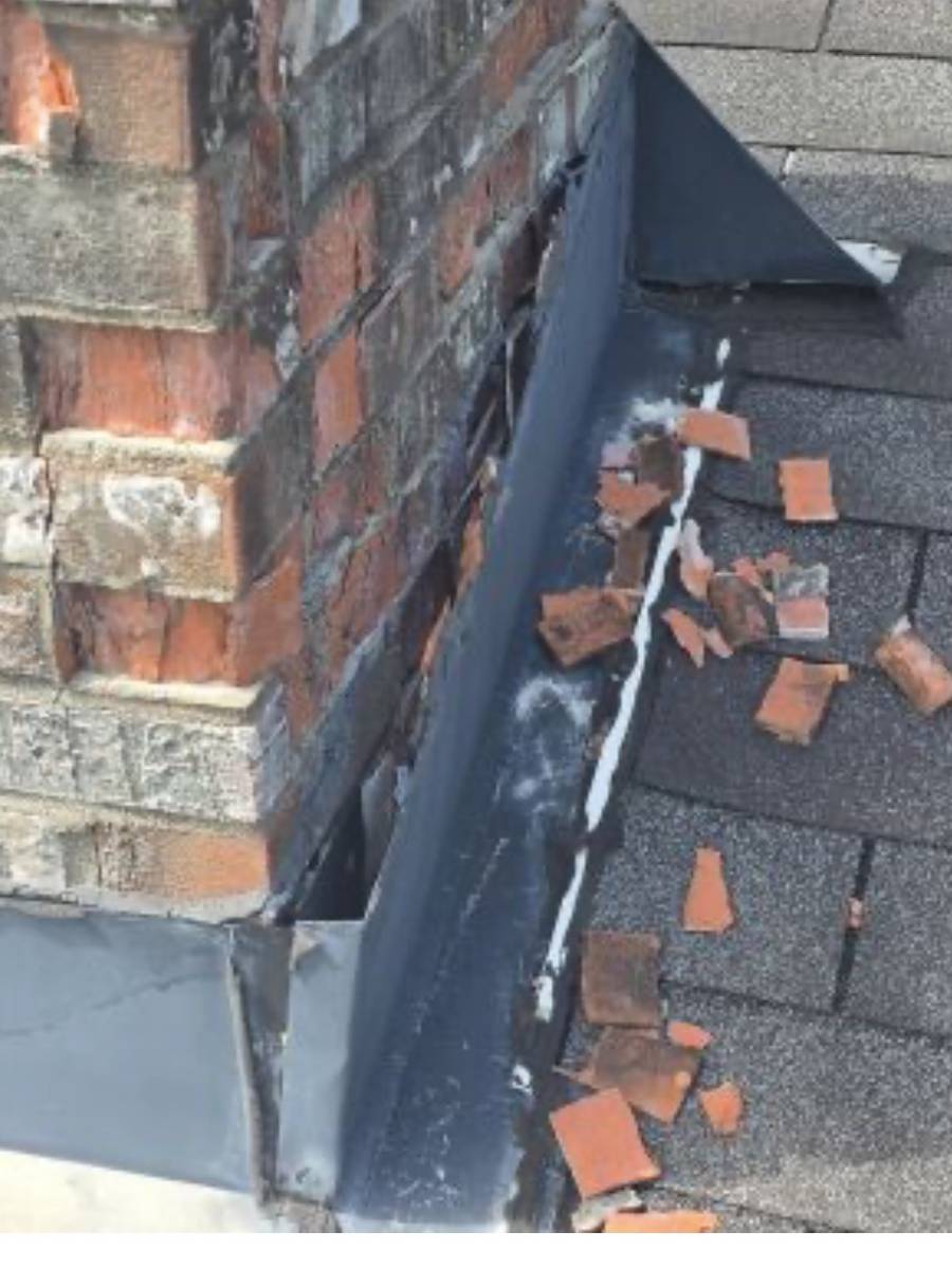 Close-up of a brick chimney meeting a shingle roof, with new dark flashing and fresh white sealant along the joint and broken tile pieces on the shingles.